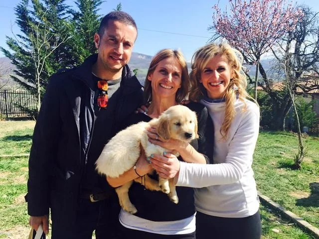 Tre persone sorridenti con un cucciolo di cane seduti in un giardino con alberi in fiore sullo sfondo.