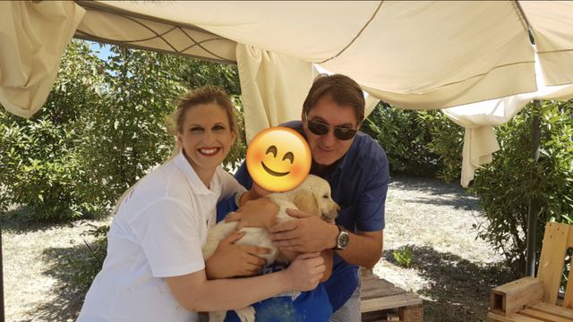 Due persone sorridenti con un cane, sotto un tendone all'aperto, in un ambiente naturale con alberi e panca di legno.