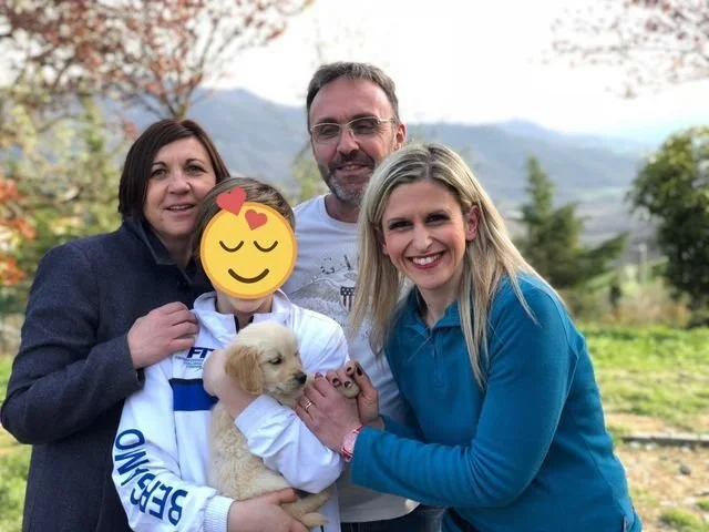 Una famiglia di quattro persone con un cane di famiglia in un paesaggio naturale con alberi e montagne sullo sfondo.