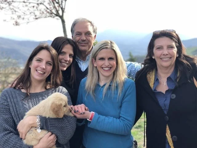 Cinque persone sorridenti, tra cui una bambina con un cucciolo di cane, all'aperto con paesaggio collinare sullo sfondo.