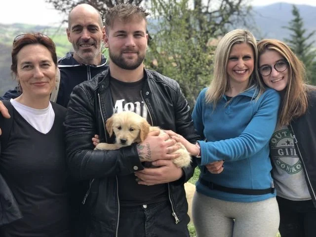 Cinque persone sorridenti e felici all'aperto con un cane (cucciolo) tra di loro, in un contesto naturale con alberi e montagne sullo sfondo.