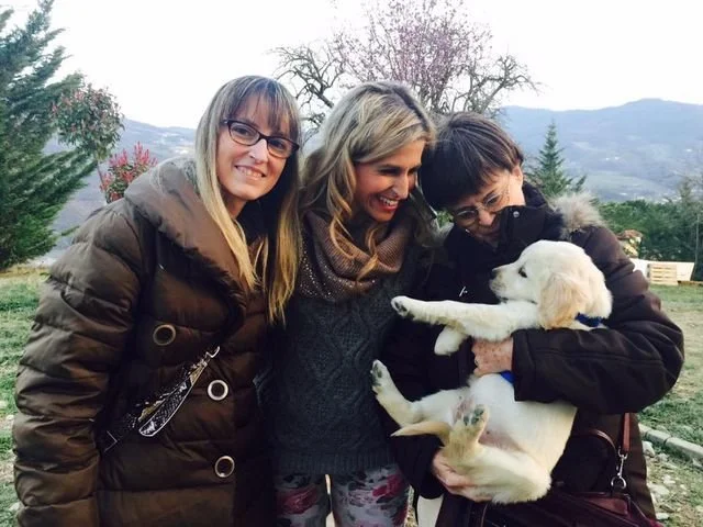 Tre donne sorridenti con un cucciolo di cane bianco in un giardino con alberi e montagne sullo sfondo.