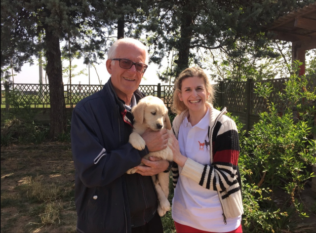 Una coppia sorridente con un cane da cucciolo in un giardino soleggiato, con alberi e recinzioni sullo sfondo.