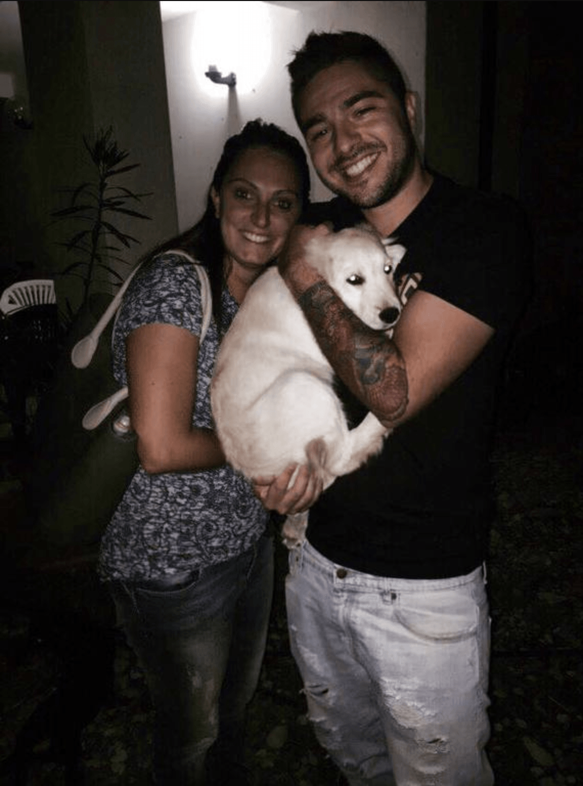 Una coppia sorridente tiene in braccio un cucciolo di cane bianco e nero, in un ambiente al chiuso, con luci soffuse.