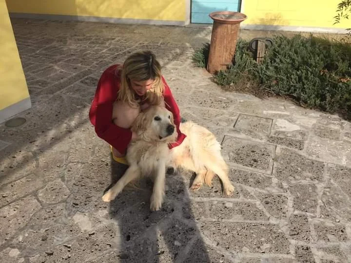 Donna che abbraccia un cane grande in una corte di pietra, con pareti colorate e vegetazione sullo sfondo.
