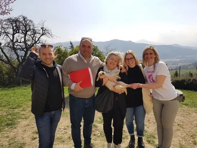 Gruppo di cinque persone sorridenti all'aperto con un cane bianco, sfondo di monti e cielo nuvoloso.