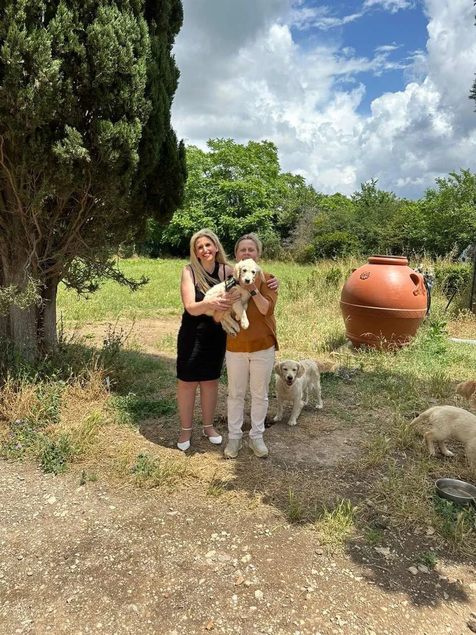 Due donne sorridenti che tengono un cane cucciolo in un giardino soleggiato, con due altri cani e un grande vaso di terracotta sullo sfondo.