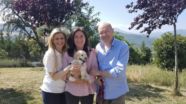 Tre persone sorridenti con un cane piccolo, in un parco verde con alberi e montagne sullo sfondo.