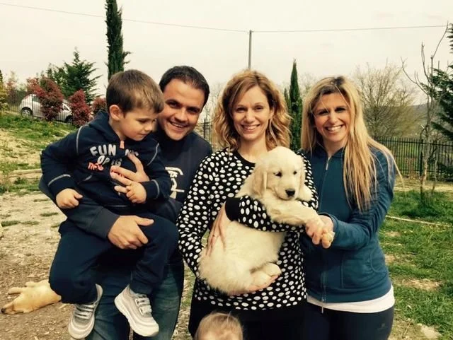 Quattro persone sorridenti e un cucciolo di cane in un giardino con alberi e recinzione.