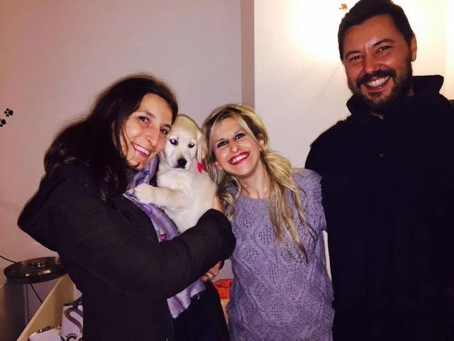 Tre persone sorridenti e una cagnolina bianca con occhi scuri in un ambiente interno.