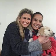 Due donne sorridenti che abbracciano un cane bianco con un collare rosso, in un ambiente interno.