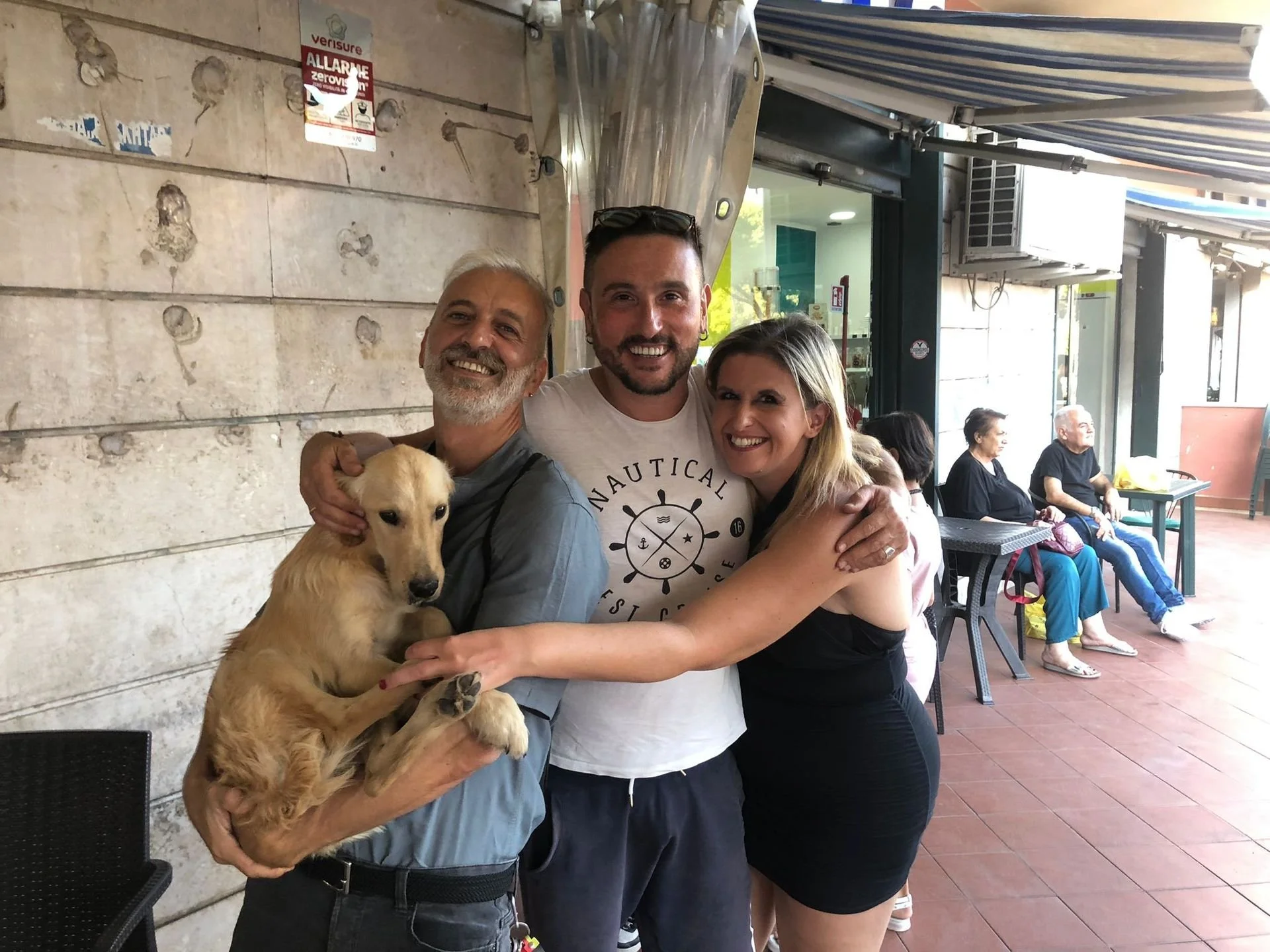 Tre persone felici abbracciate, una delle quali tiene un cucciolo di cane dorato in braccio, all'interno di un locale con parete di pietra e altre persone sedute sullo sfondo.
