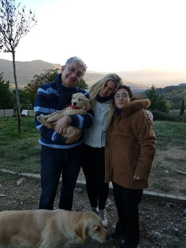Tre persone sorridenti con due cani in un parco con paesaggi collinari sullo sfondo, durante il tramonto.