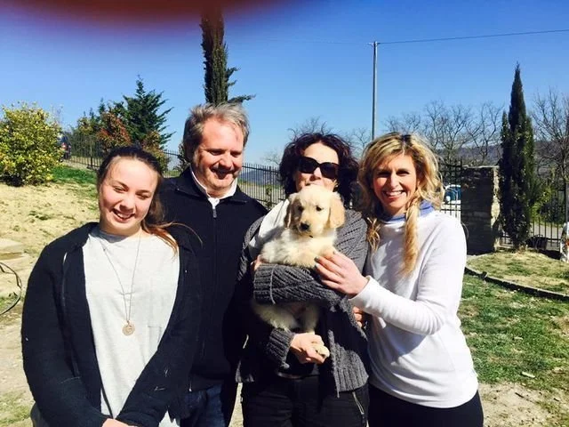Quattro persone sorridenti con un cane da cucciolo in un giardino soleggiato.