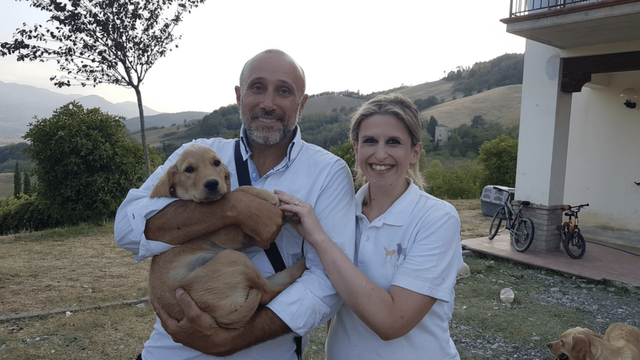 Due persone sorridenti, un uomo e una donna, con un cane giallo. Sono all'aperto con colline e alberi sullo sfondo.