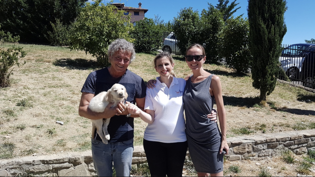 Tre persone sorridenti con un cane bianco in un'area all'aperto con alberi e cielo azzurro.