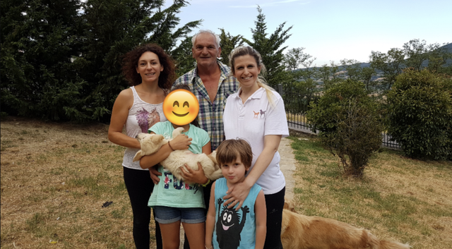 Una famiglia di cinque persone e un cane in un giardino con alberi e cespugli.