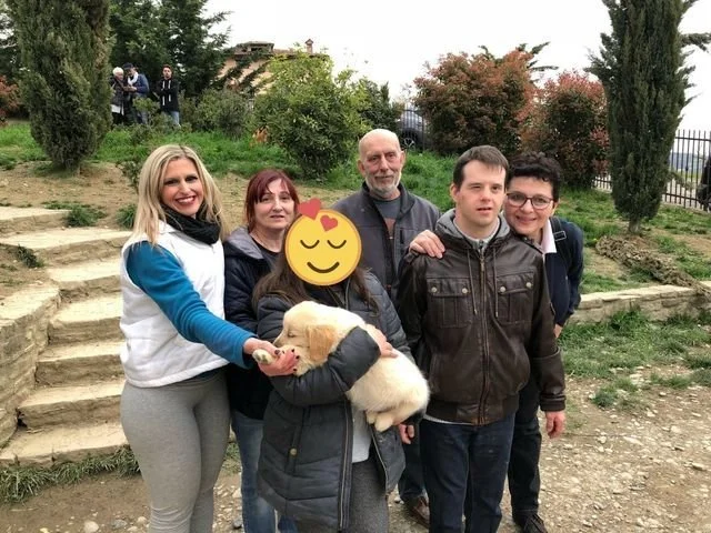 Gruppo di sei persone che posano all'aperto, alcune sorridono e una tiene un cucciolo di cane, in un ambiente naturale con alberi e scale in pietra.