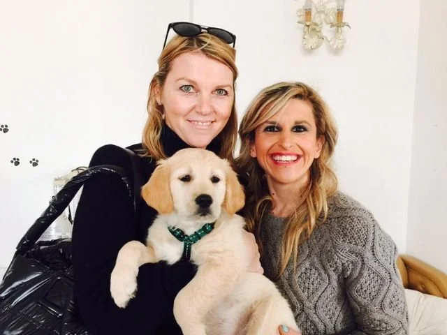 Due donne sorridenti con un cane labrador giallo, in un ambiente casalingo.