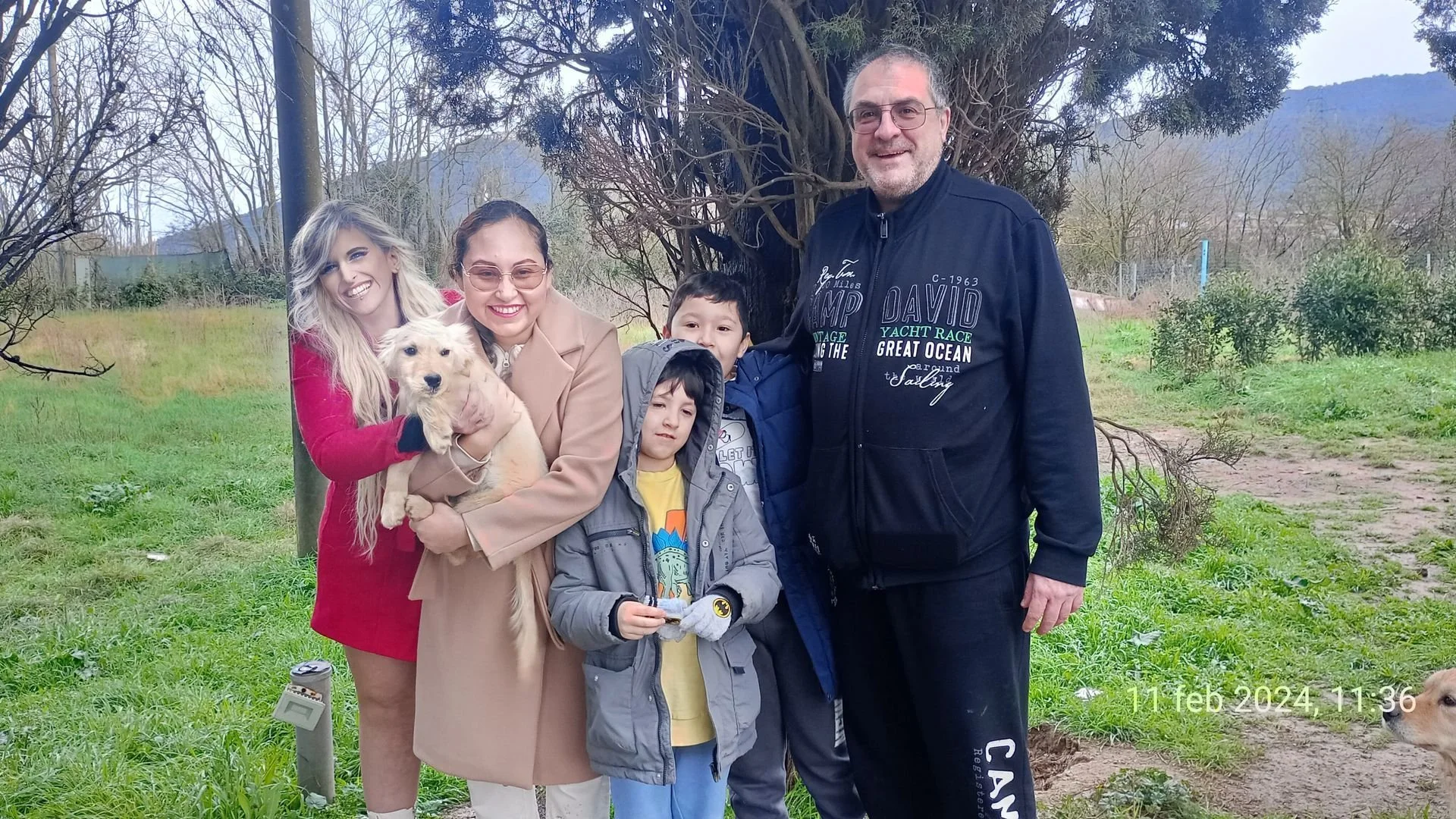 Gruppo di sei persone, adulti e bambini, sorridenti, all'aperto in un parco con alberi spogli, alcuni con cappotti, e un cane bianco con macchie marroni, sotto un albero. Data e ora visualizzate in basso a destra: 11 febbraio 2024, 11:36.