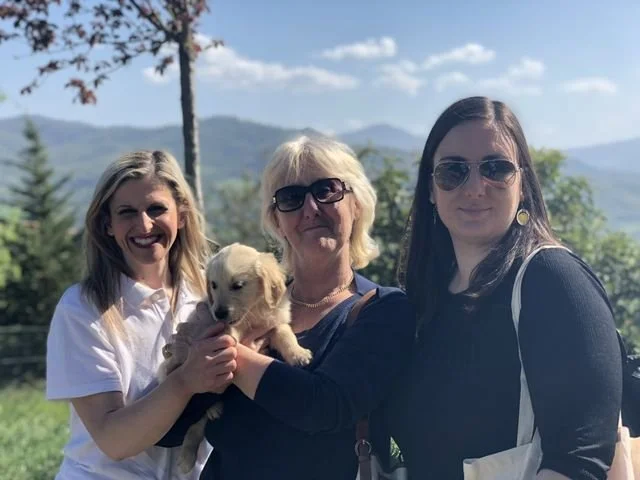 Tre donne sorridenti all'aperto, una tiene un cucciolo di cane, sfondo di natura e montagne.