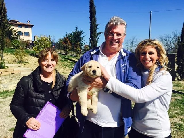Tre persone sorridenti in giardino con un cane cucciolo, uno con un foglio viola in mano.