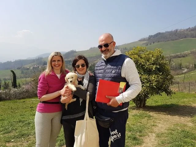 Tre persone sorridenti con un cane da compagnia in un paesaggio collinare verde, con un uomo che tiene dei documenti e una donna con occhiali.