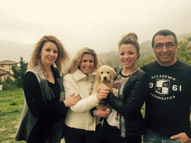 Quattro persone e un cagnolino in un paesaggio rurale con colline e alberi, in una giornata nebbiosa