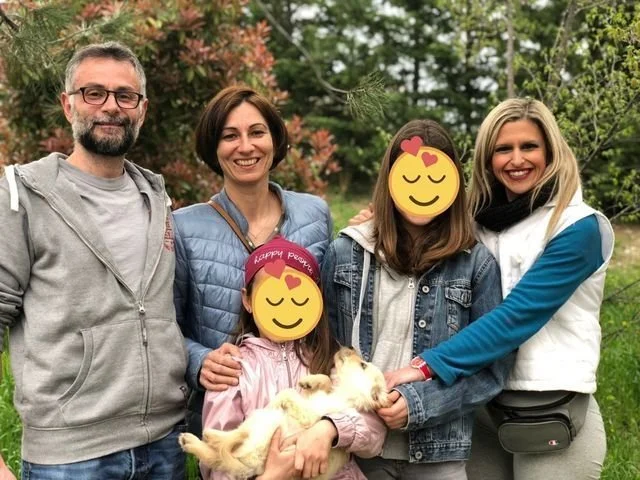 Cinque persone che sorridono all'aperto con un cane di pezza, in un parco con alberi verdi e fiori.
