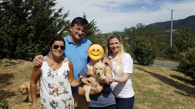 Famiglia di quattro persone con due cuccioli di cane in un giardino soleggiato, con alberi e colline sullo sfondo.