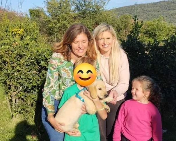 Tre donne, due bambine e un cane golden retriever in giardino, sorridenti e felici, con sfondo di natura e montagne.