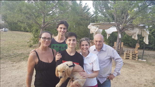 Cinque persone sorridenti e un cucciolo di cane sono riuniti all'aperto in un'area di campagna con alberi e una tende con sedie di legno sullo sfondo.