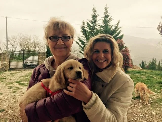 Due donne sorridenti, una anziana e una più giovane, tengono un cane di pelo chiaro, cucciolo, all'aperto in un'area verde, con un cancello e alberi sullo sfondo.