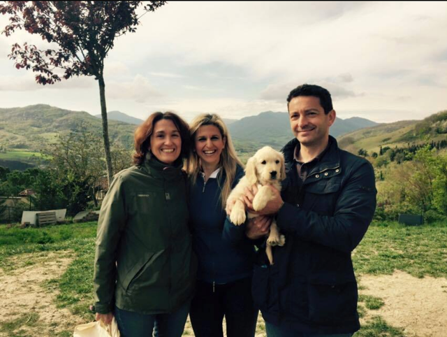 Tre persone sorridenti con un cane da compagnia in un paesaggio naturale con montagne e cielo nuvoloso sullo sfondo.
