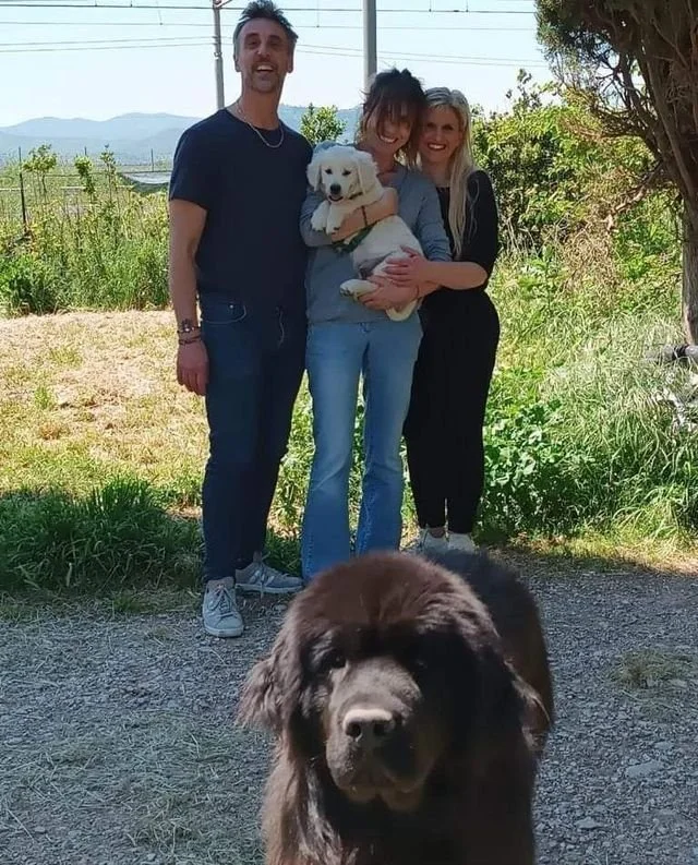 Tre persone sorridenti con due cani, uno bianco e uno nero, all'aperto in un ambiente naturale con alberi e cielo.