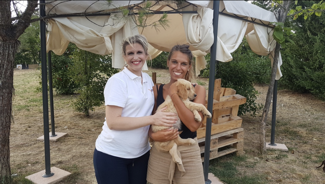 Due donne sorridenti con un cane giallo in un giardino con gazebo e panchine di legno.