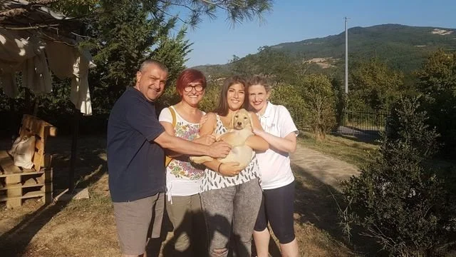 Quattro persone con un cane in un ambiente all'aperto con alberi e montagne sullo sfondo.