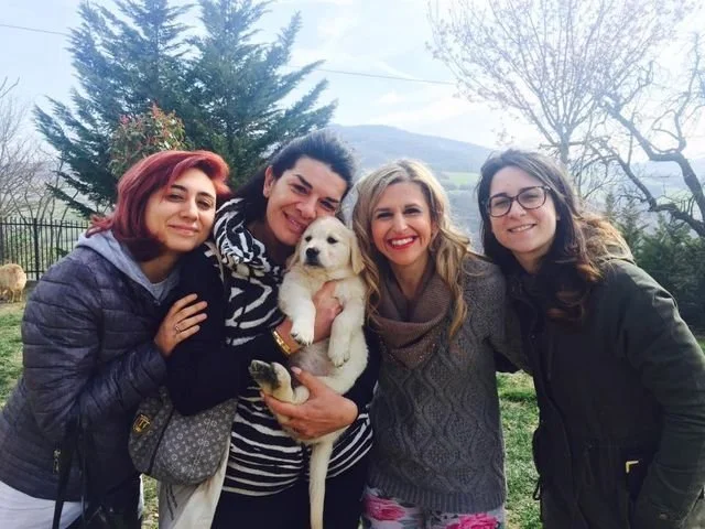 Cinque donne sorridenti con un cane bianco in un ambiente naturale.