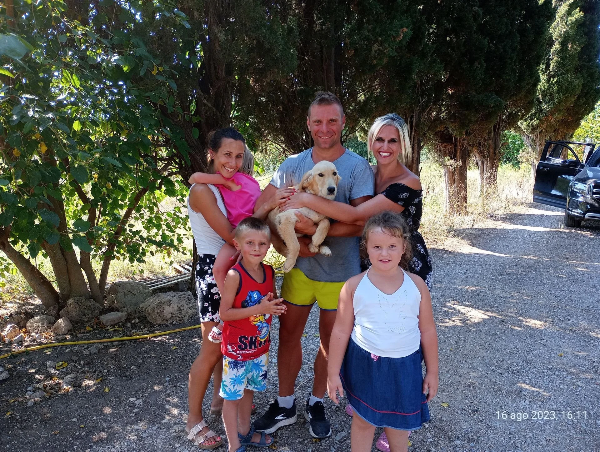 Un gruppo di persone, adulti e bambini, posano con un cane da compagnia su una strada sterrata circondata da alberi e vegetazione. È una giornata soleggiata a agosto.