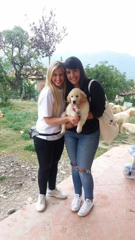 Due donne sorridenti, una con i capelli biondi e l'altra con i capelli neri, tengono un cucciolo di cane da compagnia tra di loro, in un'area all'aperto con alberi e alcuni cani sullo sfondo.
