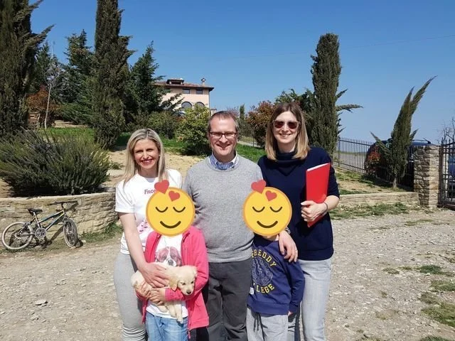 Quattro persone, due adulti e due bambini, sorridenti, stanno all'aperto in un parco, con un cane piccolo in braccio a una bambina, e un uomo e una donna sorridenti vicino a loro, sotto un cielo sereno e con alberi sullo sfondo.