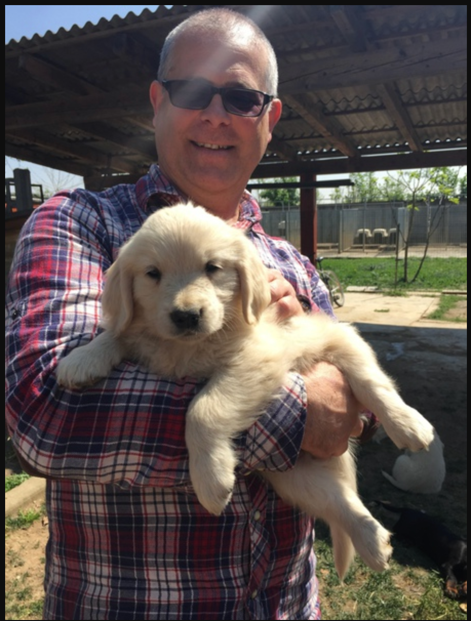 Un uomo con capelli corti e occhiali da sole sorride mentre tiene in braccio un cucciolo di golden retriever.