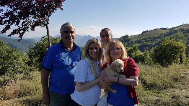 Quattro persone sorridenti con un cucciolo di cane in un paesaggio collinare verde.