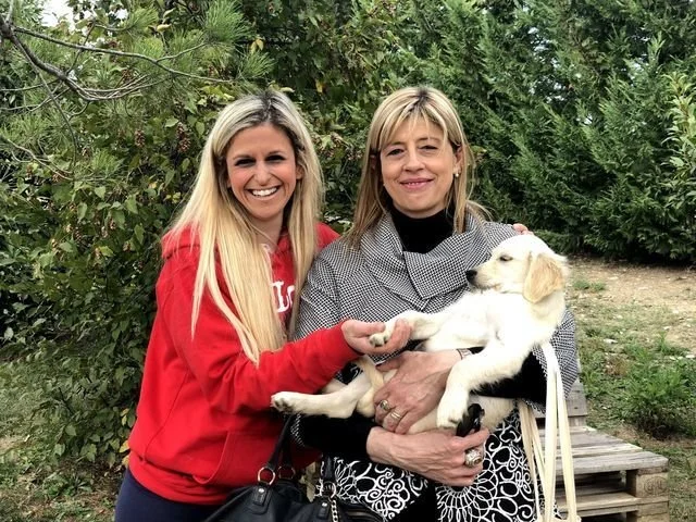Due donne sorridenti, una giovane e una più anziana, tengono un cucciolo di cane bianco e giallo in un ambiente esterno con alberi e vegetazione.