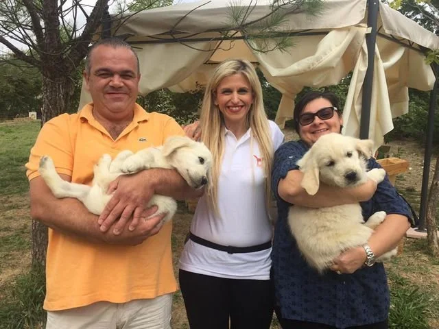 Tre persone sorridenti con due cuccioli di cane, in un giardino con gazebo.