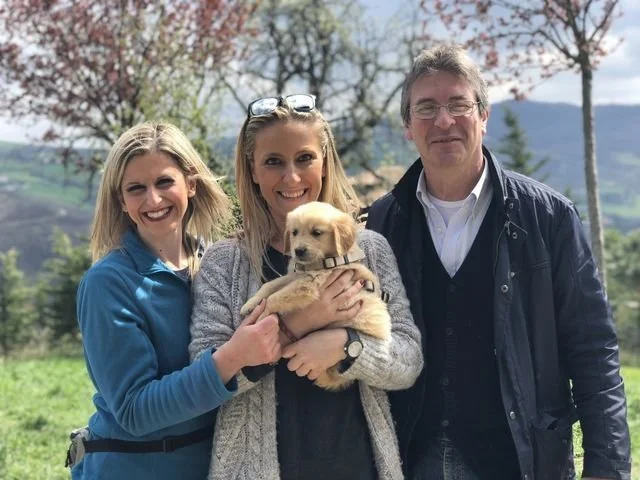 Tre persone sorridenti che tengono un cucciolo di cane, con uno sfondo di natura e alberi in fiore.