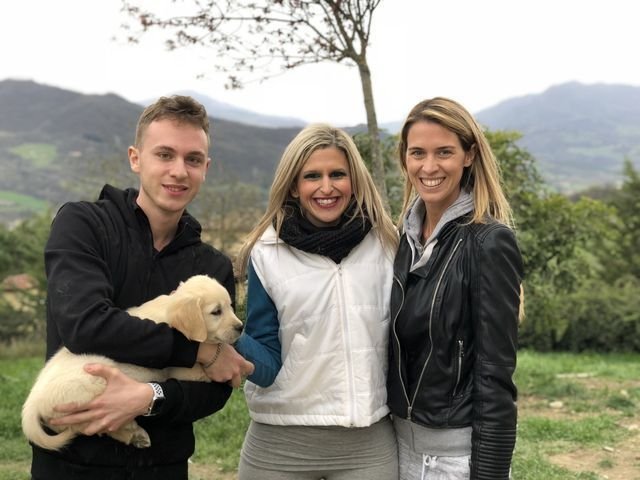 Tre donne sorridenti con un cane da compagnia in un paesaggio naturale con alberi e colline sullo sfondo.