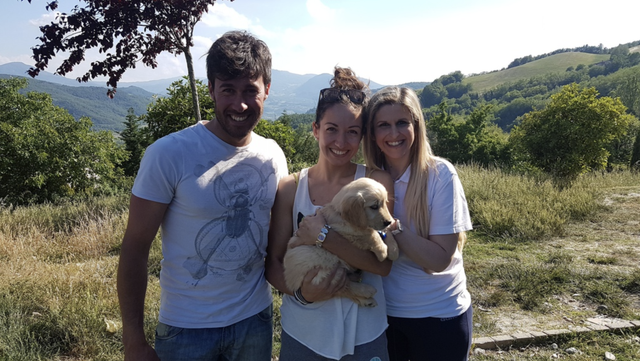 Tre persone sorridenti con un cane in un paesaggio naturale con alberi e montagne sullo sfondo.