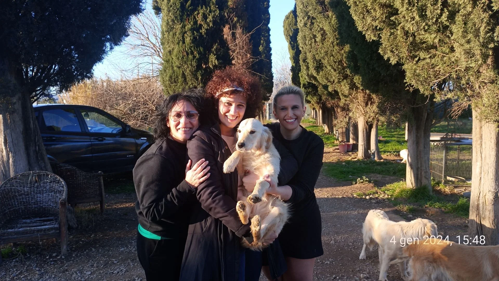 Tre donne sorridenti che tengono un cucciolo di golden retriever in un cortile con alberi e cani. È pomeriggio, 4 gennaio 2024, alle 15:48.
