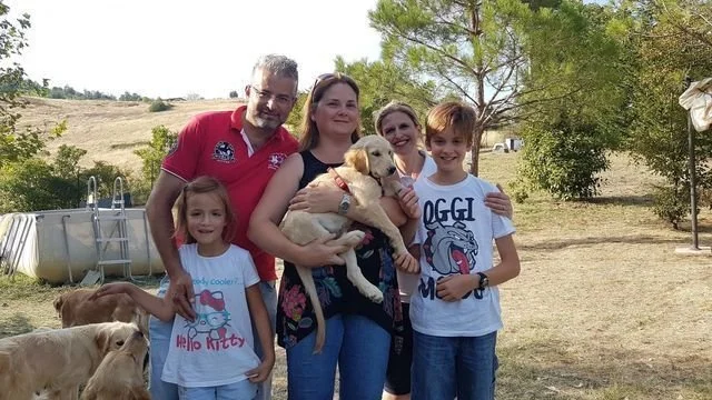 Una famiglia con bambini e cani all'aperto in un'area di campagna o parco, con alberi e un campo in background.